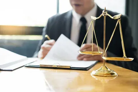 Un abogado con traje escribe en un escritorio con una balanza de justicia dorada en primer plano.