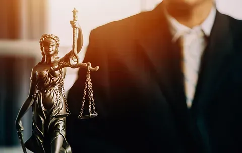 Estatua de la Dama de la Justicia con una balanza, frente a una persona con traje. Una luz cálida ilumina la escena.