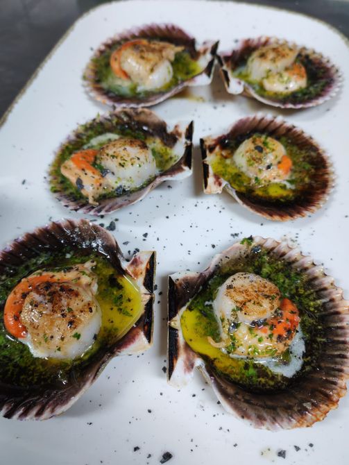 Vieiras selladas en sus conchas, rociadas con salsa verde, en un plato blanco.