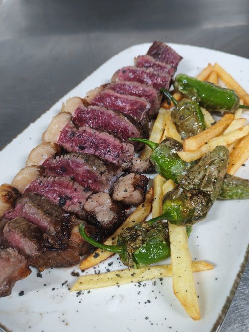 Filete en rodajas, patatas fritas y pimientos verdes a la parrilla en un plato blanco.
