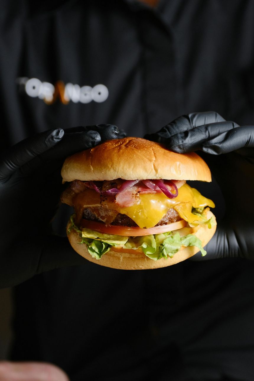 Hamburguesa sostenida por manos enguantadas de negro, con pan, queso, cebolla roja, tomate y lechuga.