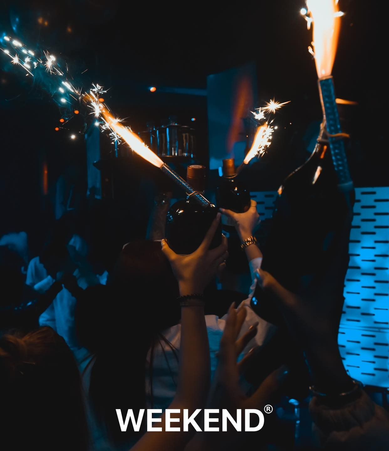 Un grupo de personas sostiene botellas de champán en sus manos con bengalas de fondo y las palabras
