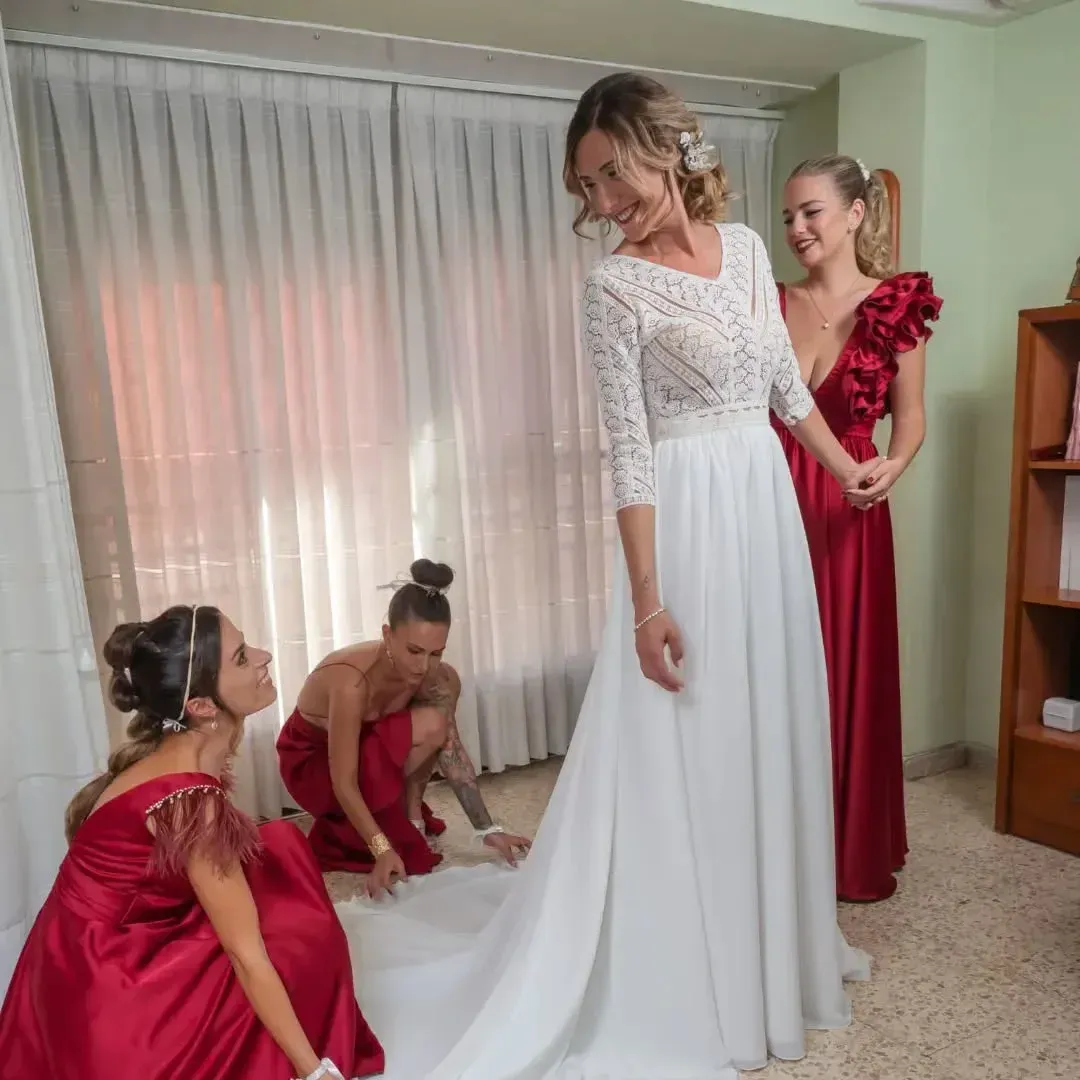La novia, vestida de blanco, sonríe mientras las damas de honor, vestidas de rojo, la ayudan. Interior con luz natural.