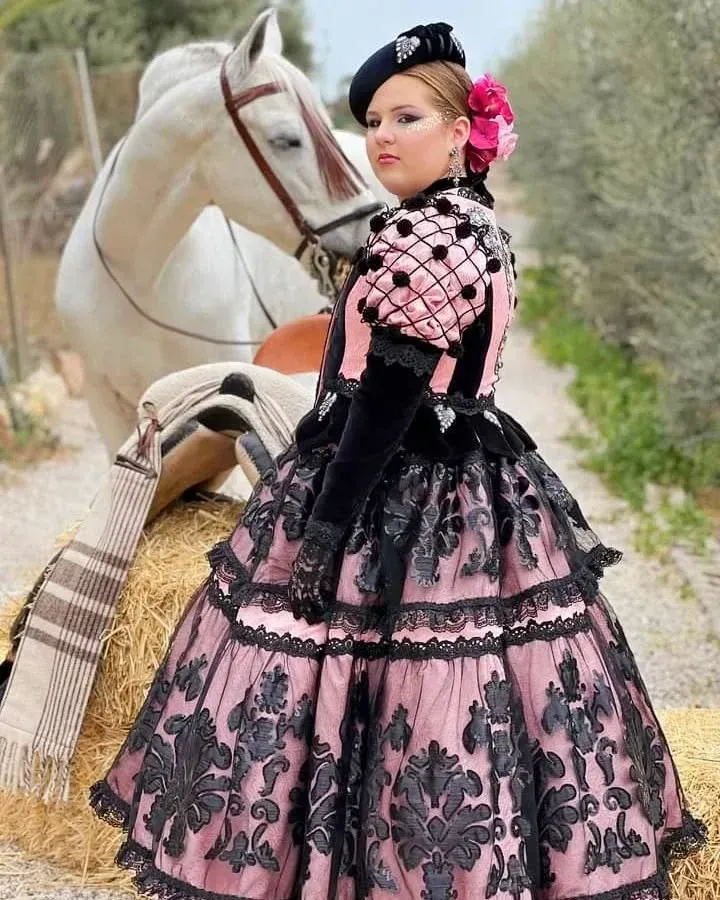 Mujer con vestido victoriano rosa y negro, de pie cerca de un caballo blanco con silla de montar en un camino.