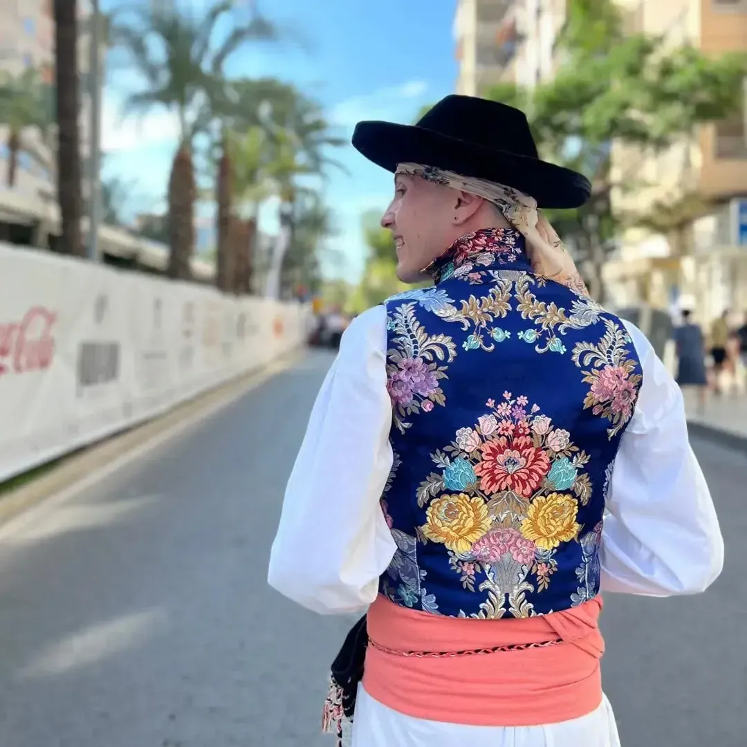 Persona con chaleco y sombrero tradicionales caminando por la calle. Colores vibrantes, bordados florales y edificios.