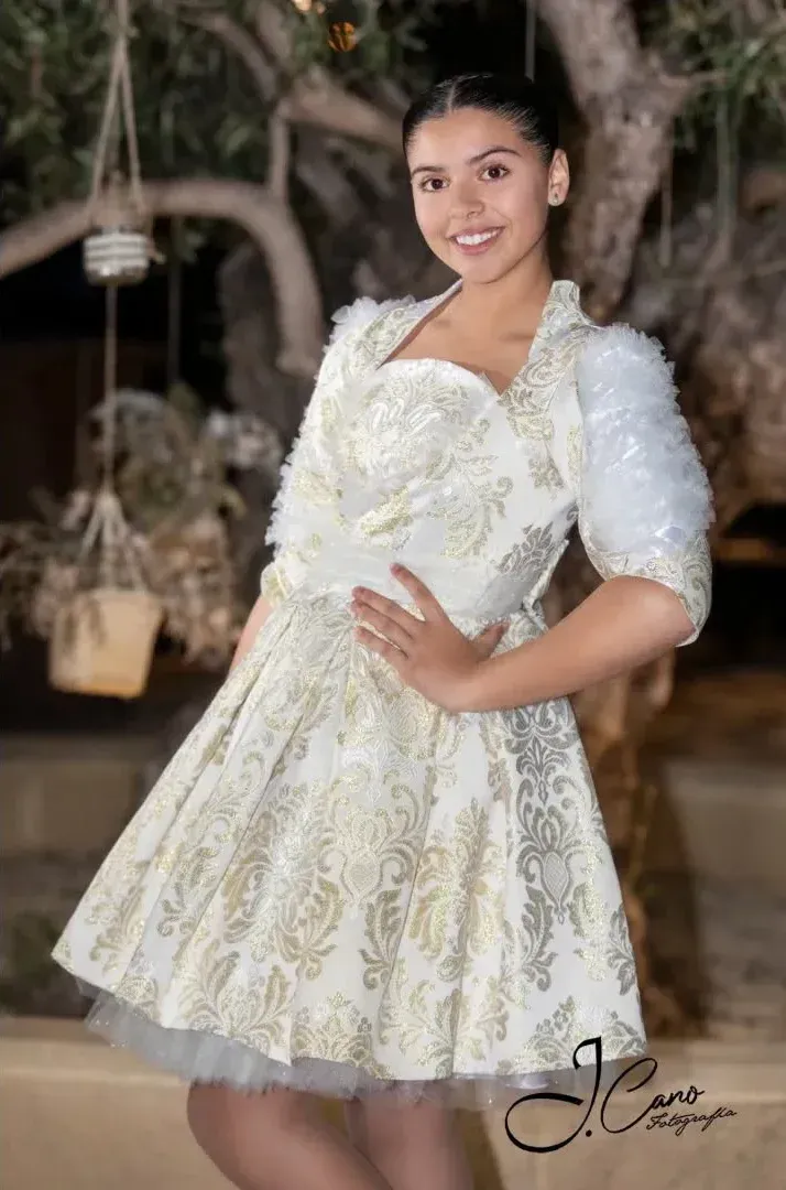 Mujer con vestido blanco y dorado estampado, mangas cortas y pelo blanco, sonriendo. Escena al aire libre.