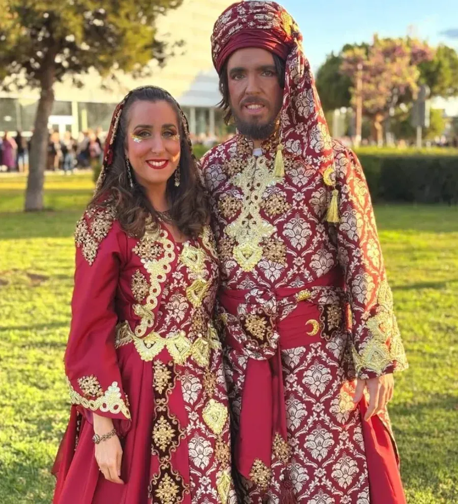Pareja vestida con ropas ornamentadas de estilo de Medio Oriente en tonos rojos y dorados, sonriendo al aire libre.