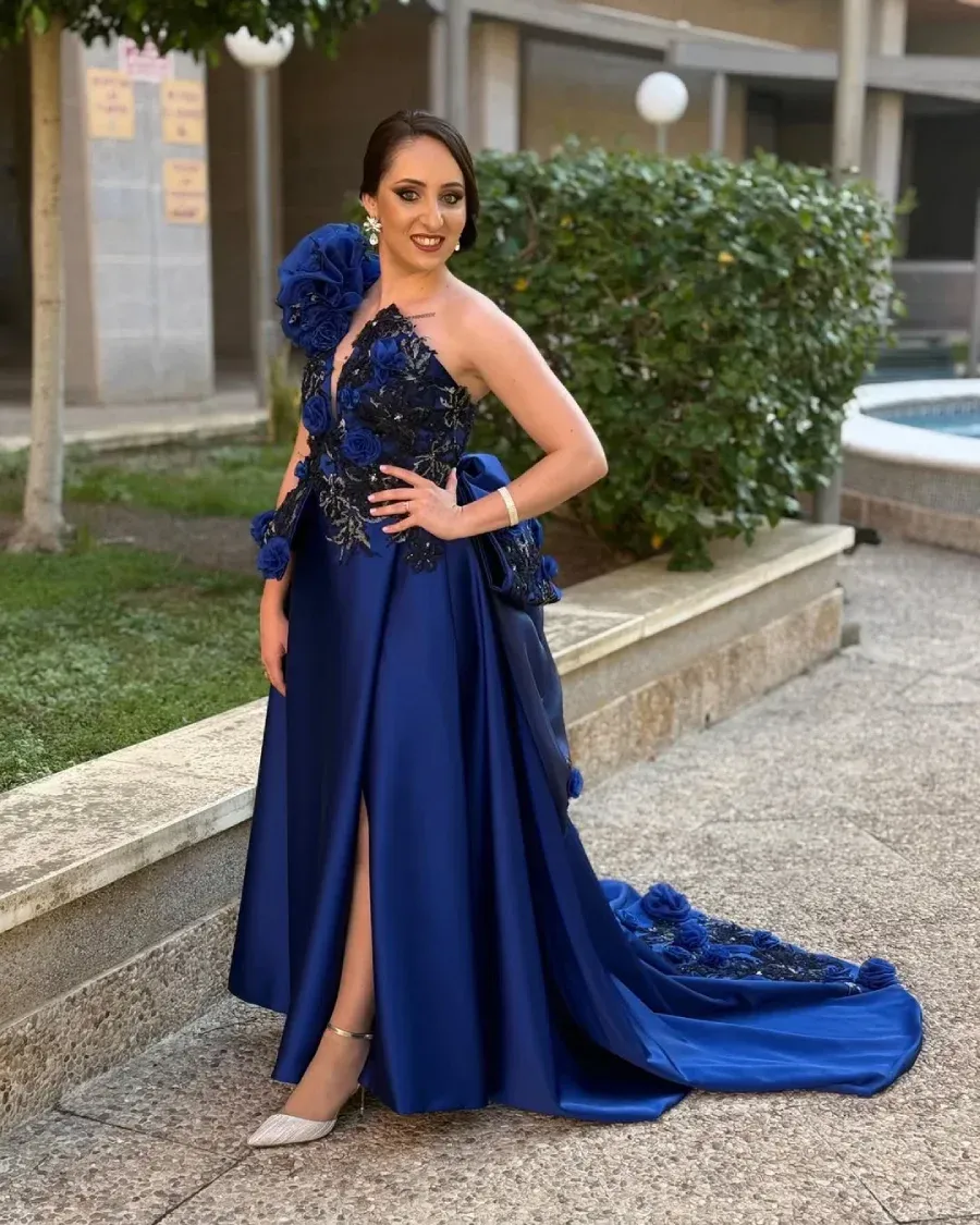 Mujer con un vestido de noche azul con detalles florales, posando al aire libre, con la mano en la cadera, sonriendo.