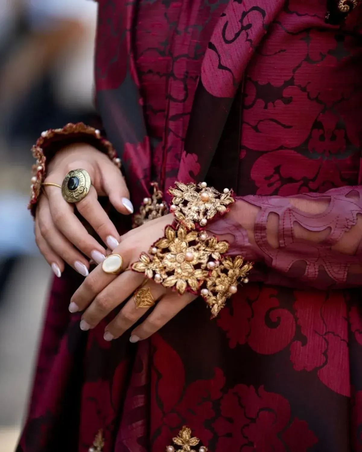 Tela con estampado floral color burdeos, joyas doradas adornadas en las manos con uñas pálidas.
