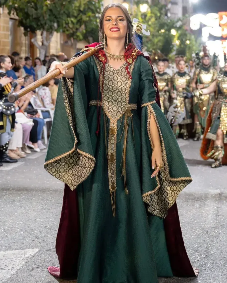 Mujer con vestido medieval verde con bastón, sonriendo en el desfile.