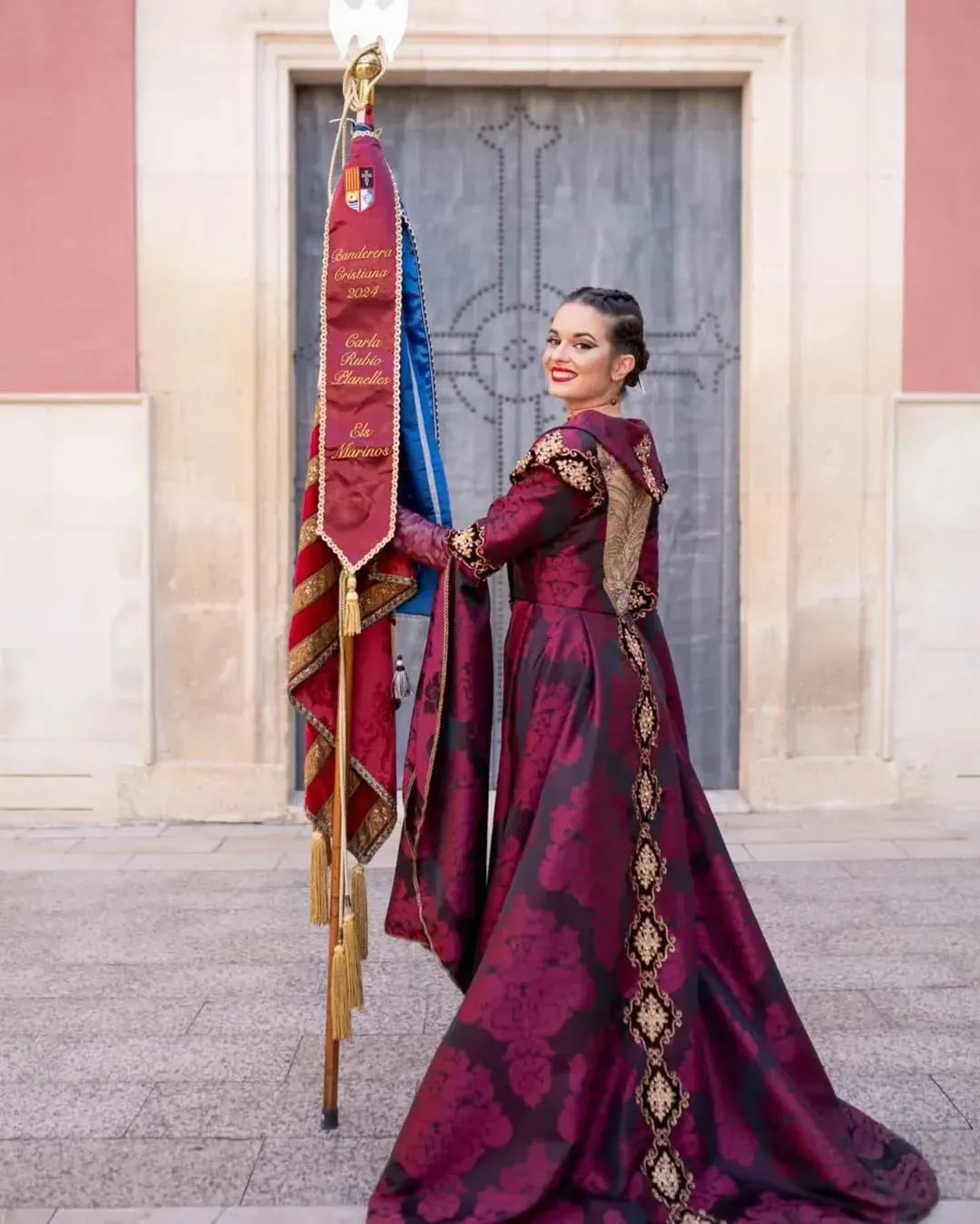 Mujer con un vestido color burdeos sosteniendo una bandera, sonriendo frente a un edificio con una gran puerta de madera.