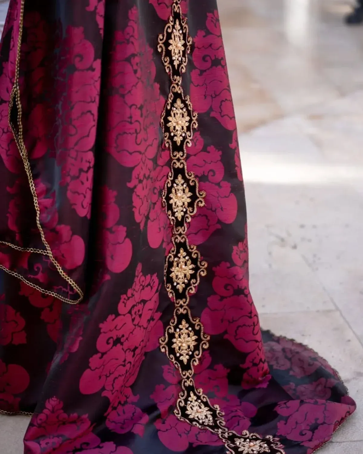 Primer plano de un vestido de damasco burdeos y negro con adornos dorados y negros a lo largo del borde.