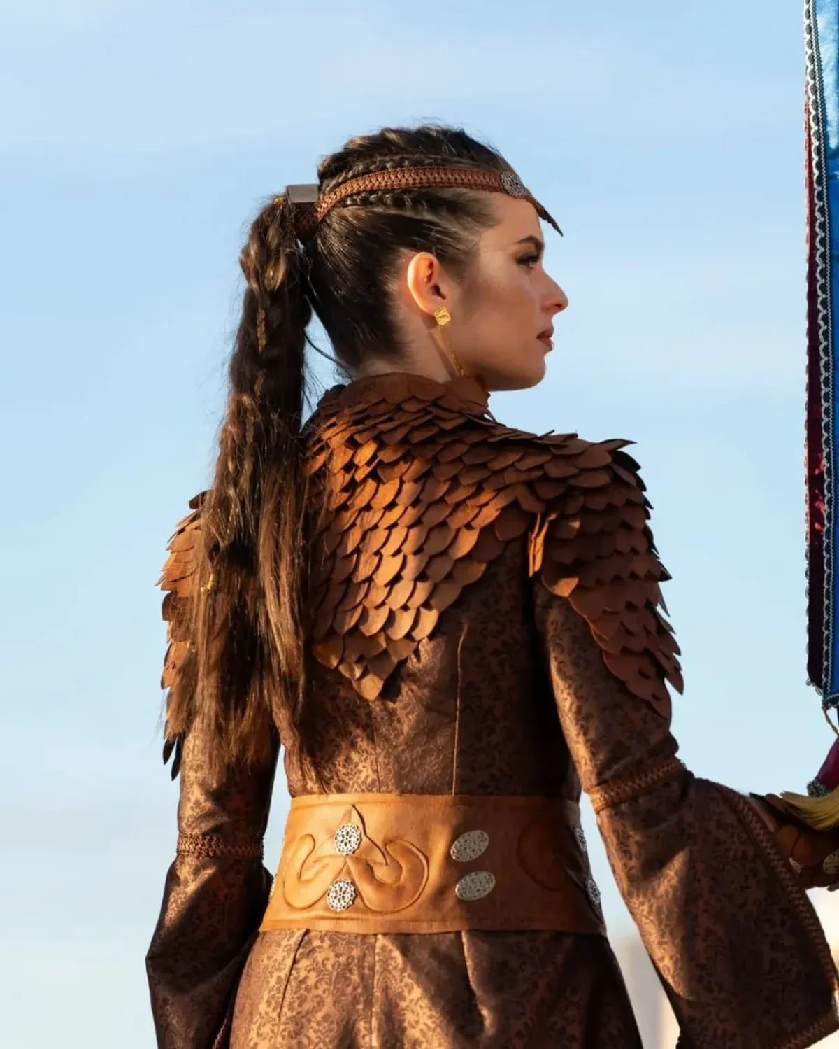 Mujer con armadura marrón, de perfil, portando un arma. Cola de caballo y diadema trenzada, al aire libre.