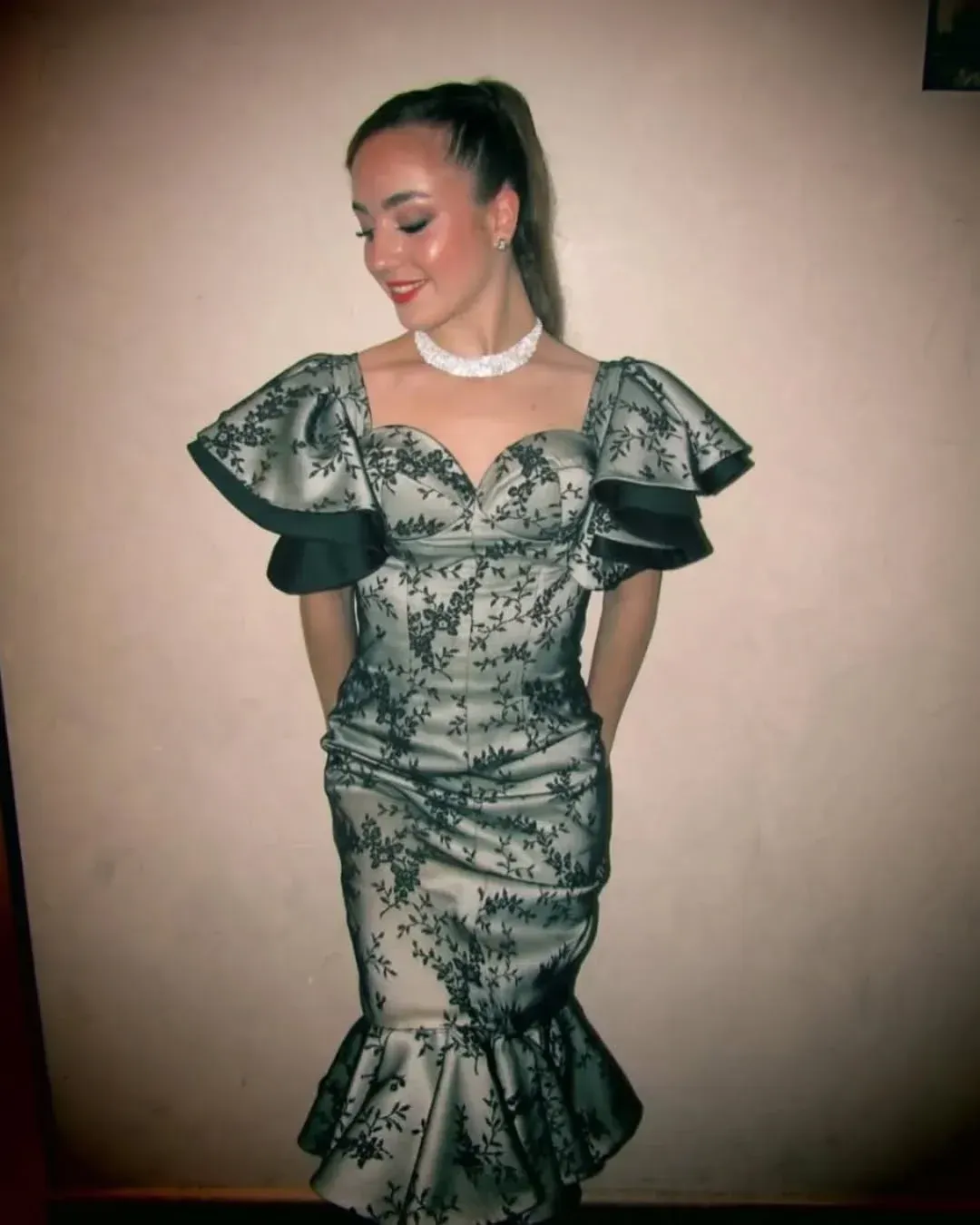 Mujer posando con vestido formal, con coleta alta y collar blanco. El vestido es negro floral sobre blanco, con mangas