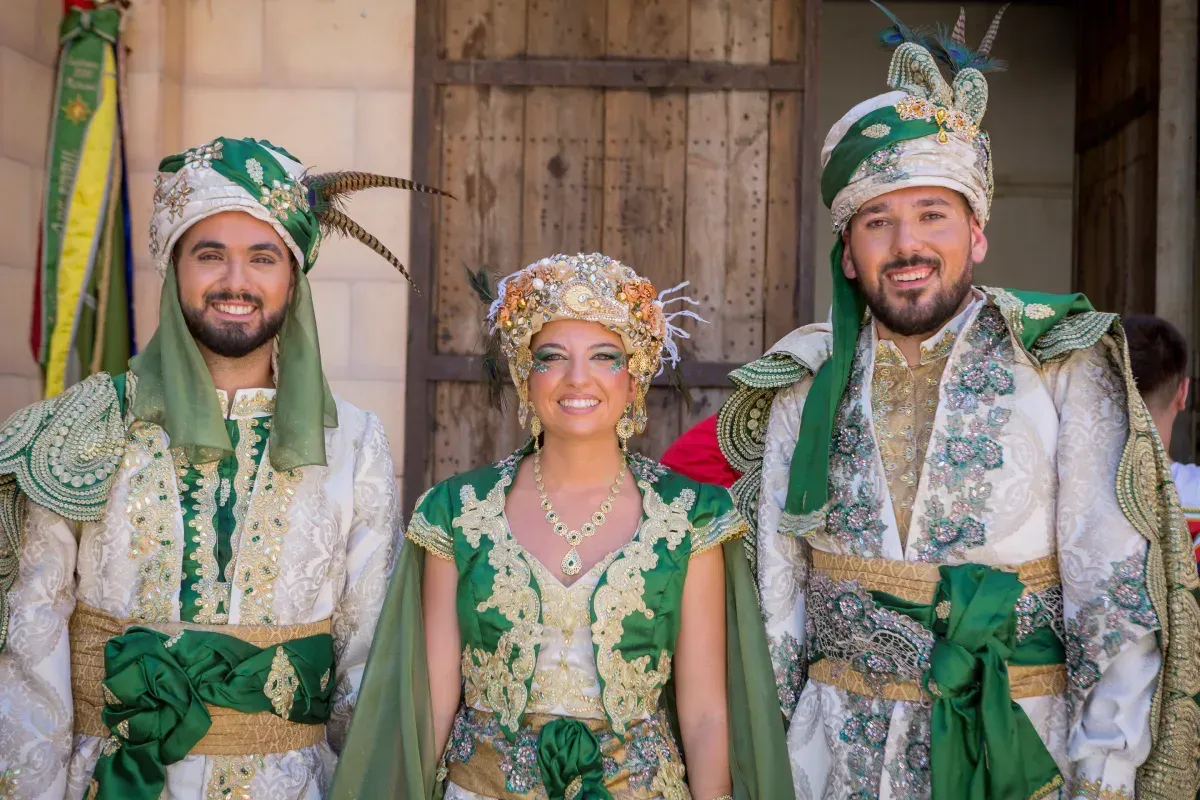 Pareja vestida con ropas ornamentadas de estilo de Medio Oriente en tonos rojos y dorados, sonriendo al aire libre.