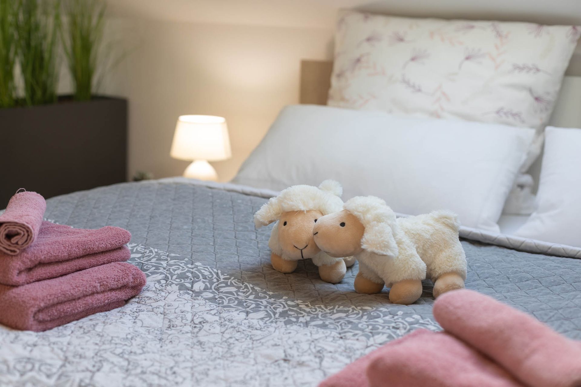 Zwei Stoffschafe sitzen auf einem Bett neben einem Stapel Handtücher.