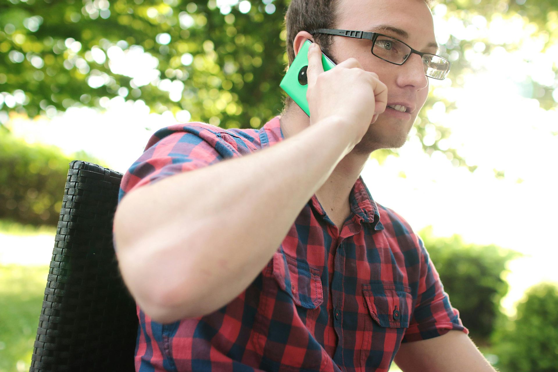 Hombre con camisa a cuadros y gafas hablando por un teléfono verde al aire libre en un jardín.