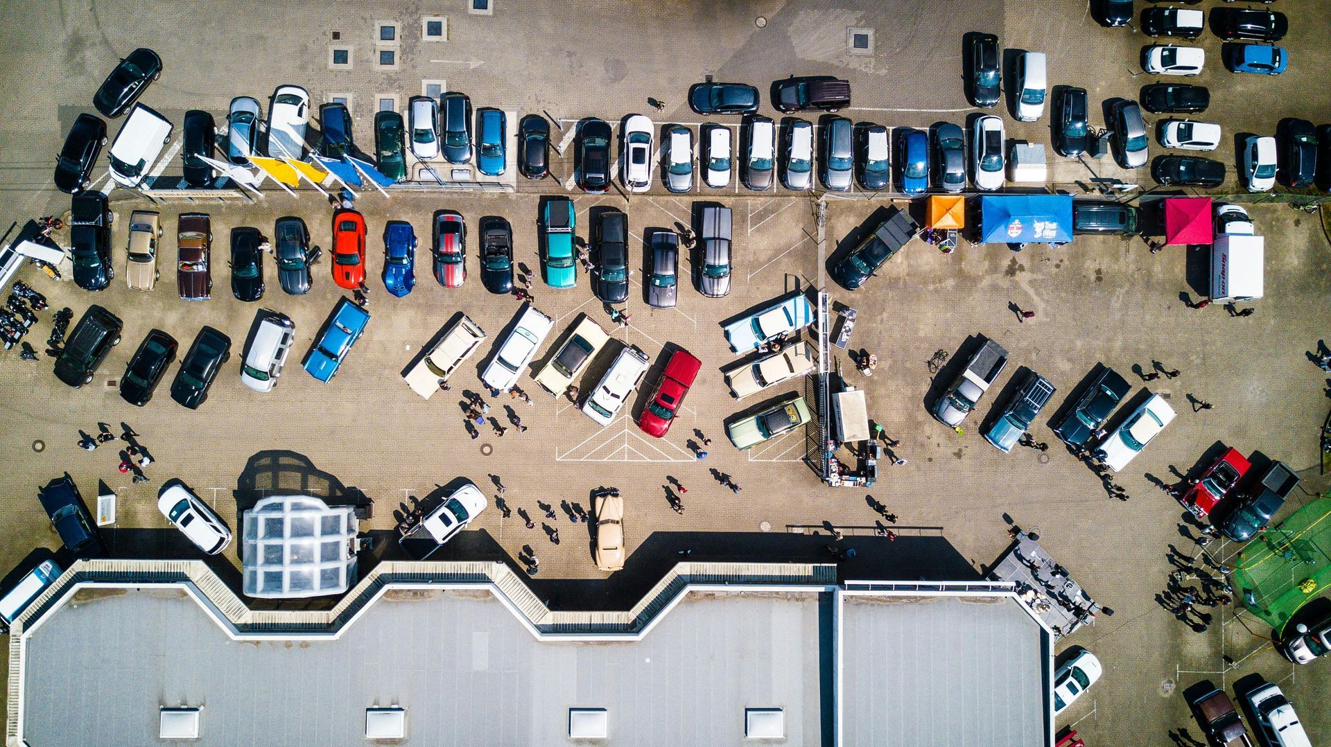 Vista aérea de un estacionamiento abarrotado de varios autos y personas reunidas alrededor de un gran edificio.
