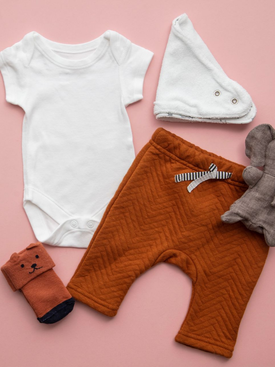 Vêtements de bébé disposés sur un fond rose : un body blanc, un bonnet crème, un pantalon rouille, des chaussettes et une peluche.