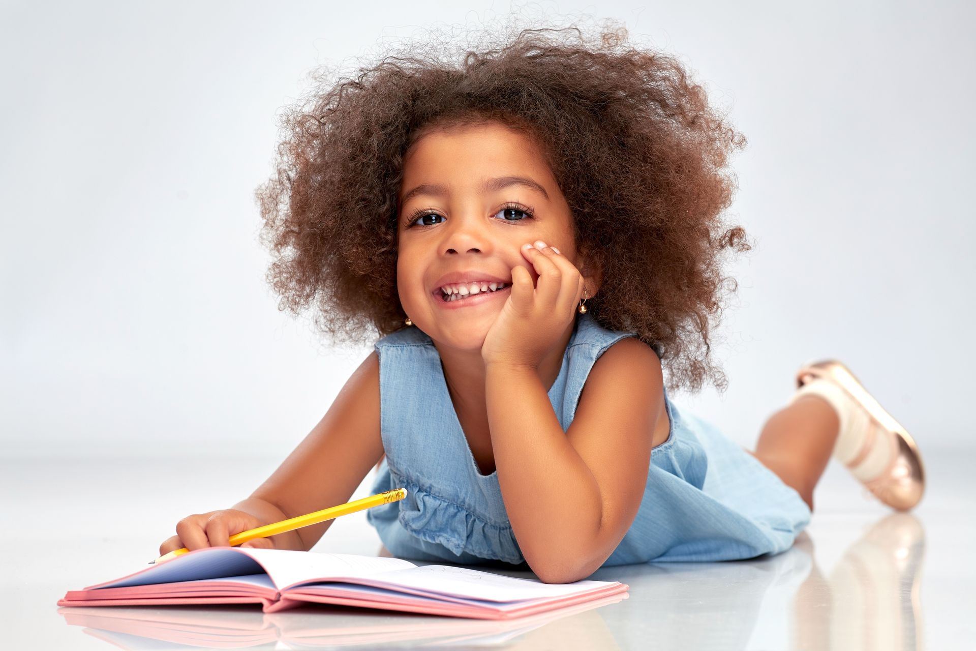 Enfant allongé sur le sol, cahier ouvert et crayon à la main, souriant à la caméra