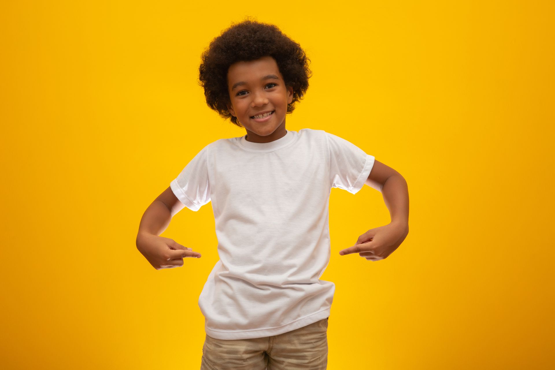 Enfant souriant en chemise blanche et pantalon beige, posant les bras écartés sur un fond jaune vif