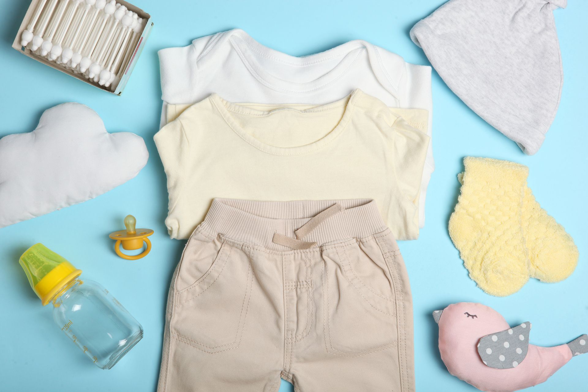 Vêtements et accessoires pour bébé disposés à plat sur un fond bleu, comprenant notamment un body, un bonnet, un bavoir, un biberon et une tétine.