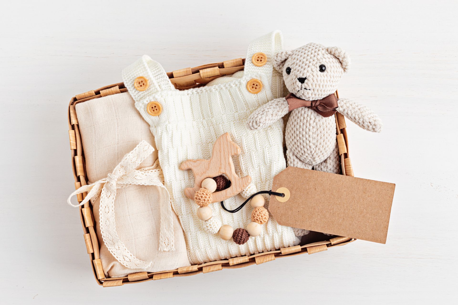 Panier-cadeau contenant un ours en peluche, une couverture blanche tricotée, des chaussettes pour bébé et une étiquette vierge, le tout présenté dans un plateau en osier.