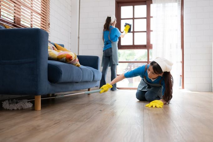 Una mujer está limpiando el piso de una sala de estar mientras otra mujer limpia una ventana.
