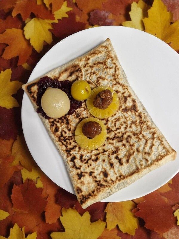 Crêpe carrée garnie, servie sur une assiette blanche, devant un décor de feuilles d'automne. Accompagnée de fruits pochés et de confiture.