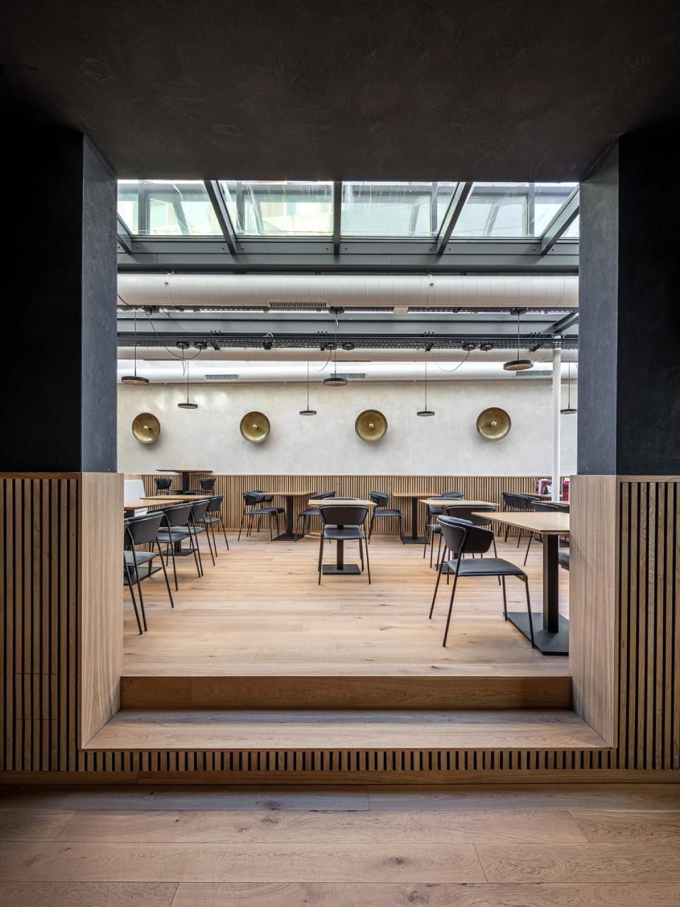 Intérieur du restaurant vu à travers une porte, avec parquet et tables en bois, lumière naturelle provenant d'une verrière.