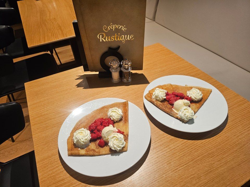 Deux crêpes à la crème et aux framboises sont présentées sur des assiettes blanches, sur une table. Un menu et des salières et poivrières sont visibles en arrière-plan.