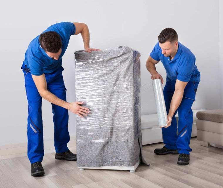 Dos operarios de mudanzas con uniformes azules envuelven un electrodoméstico blanco en film plástico en el interior.