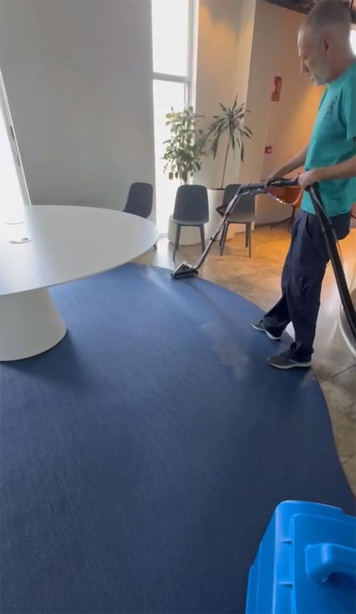 Un hombre aspira una alfombra azul en una oficina. Al fondo, una mesa redonda blanca, sillas y una planta.