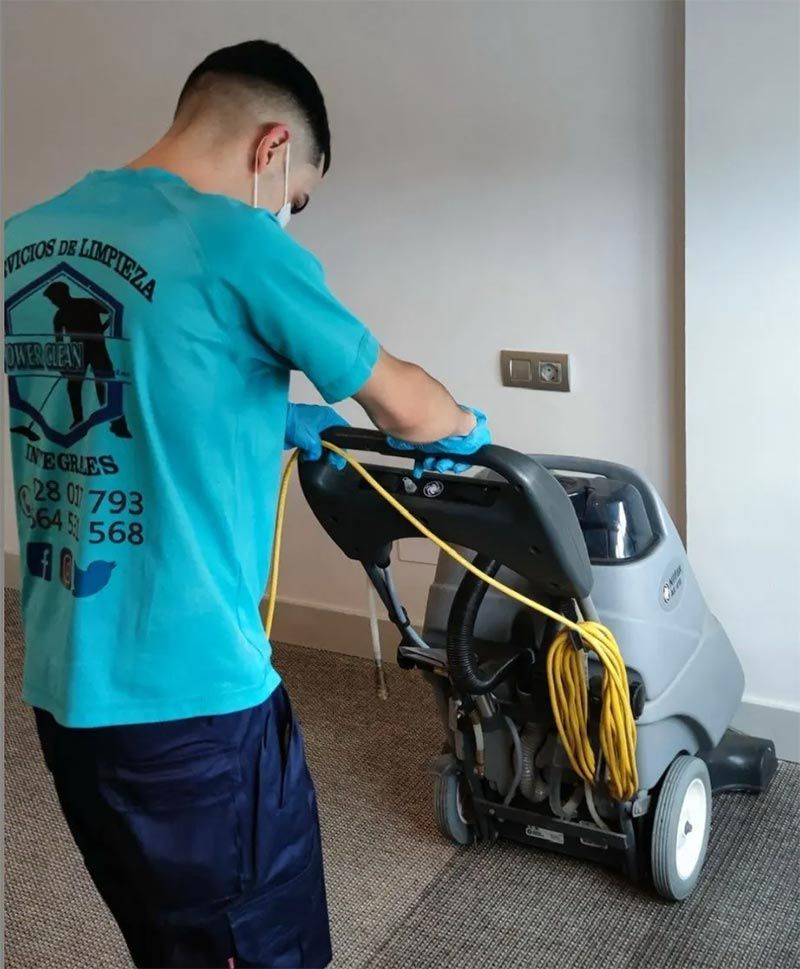 Una persona con camisa azul opera una máquina limpiadora de alfombras en un lugar interior.