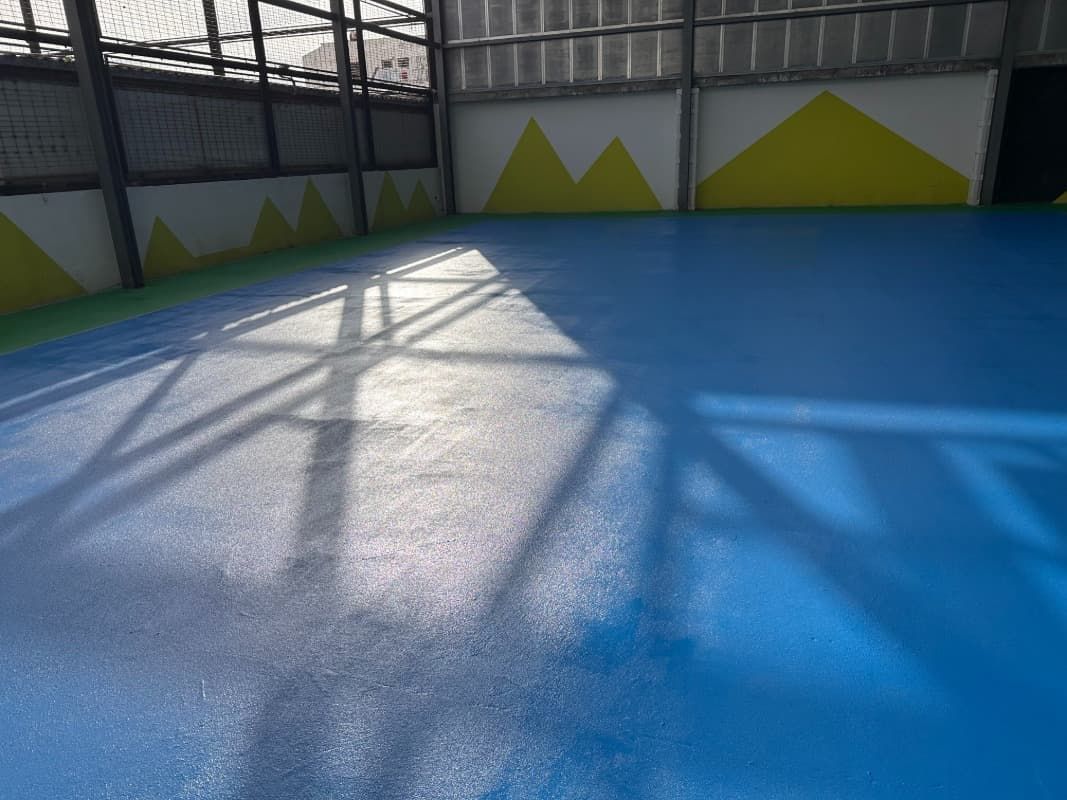 Cancha deportiva azul vacía con luz solar brillante y patrones de sombras junto a una valla y detalles de pared amarillos.