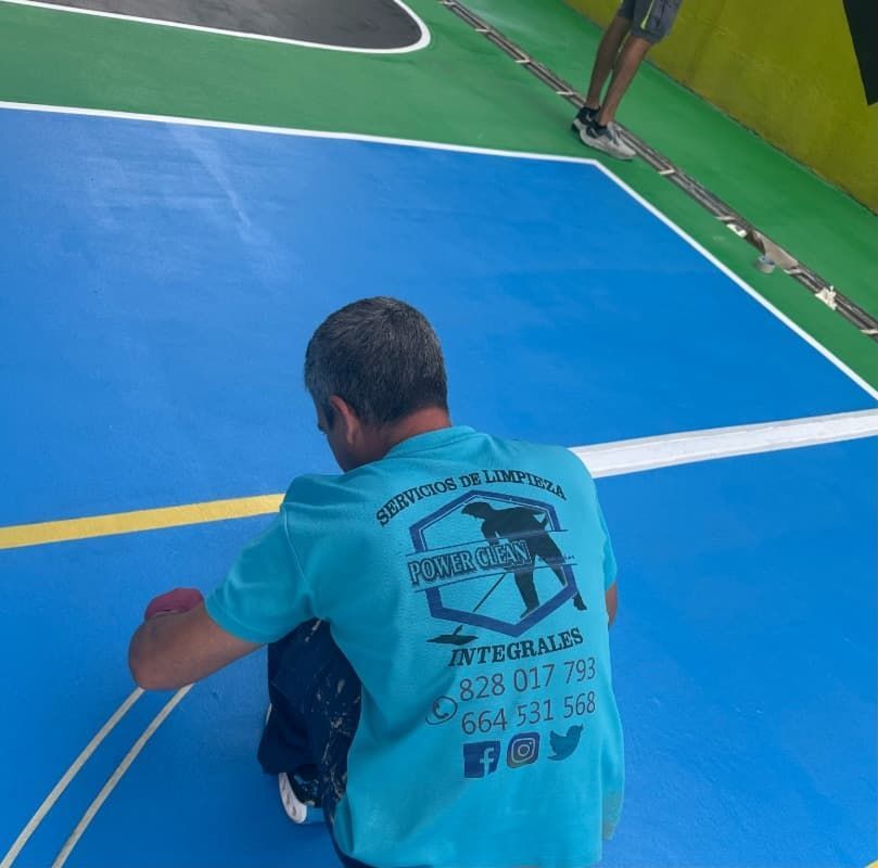 Hombre sentado en una cancha cubierta azul, visto desde atrás, con una camisa turquesa con texto impreso.