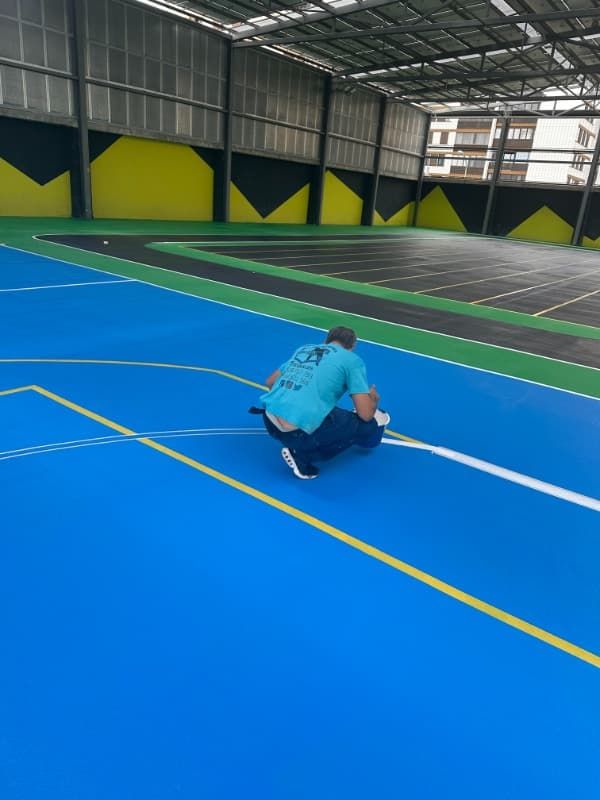 Persona agachada en una cancha deportiva cubierta azul junto a las líneas amarillas que delimitan el perímetro.