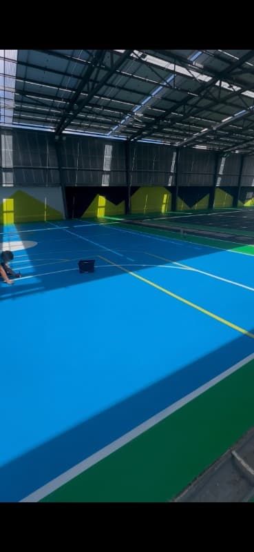 Pista de tenis cubierta de color azul y verde bajo un alto techo metálico con luces brillantes en el techo.