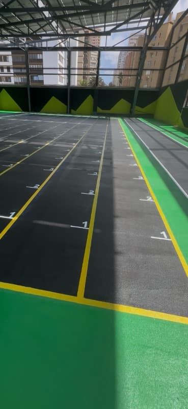 Pista deportiva cubierta con suelo oscuro, líneas amarillas y laterales verdes bajo un alto techo de cristal.