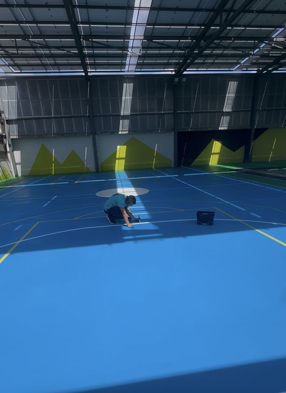 Una persona está arrodillada en una cancha de tenis cubierta azul, cerca de un cubo, en un gran pabellón deportivo.