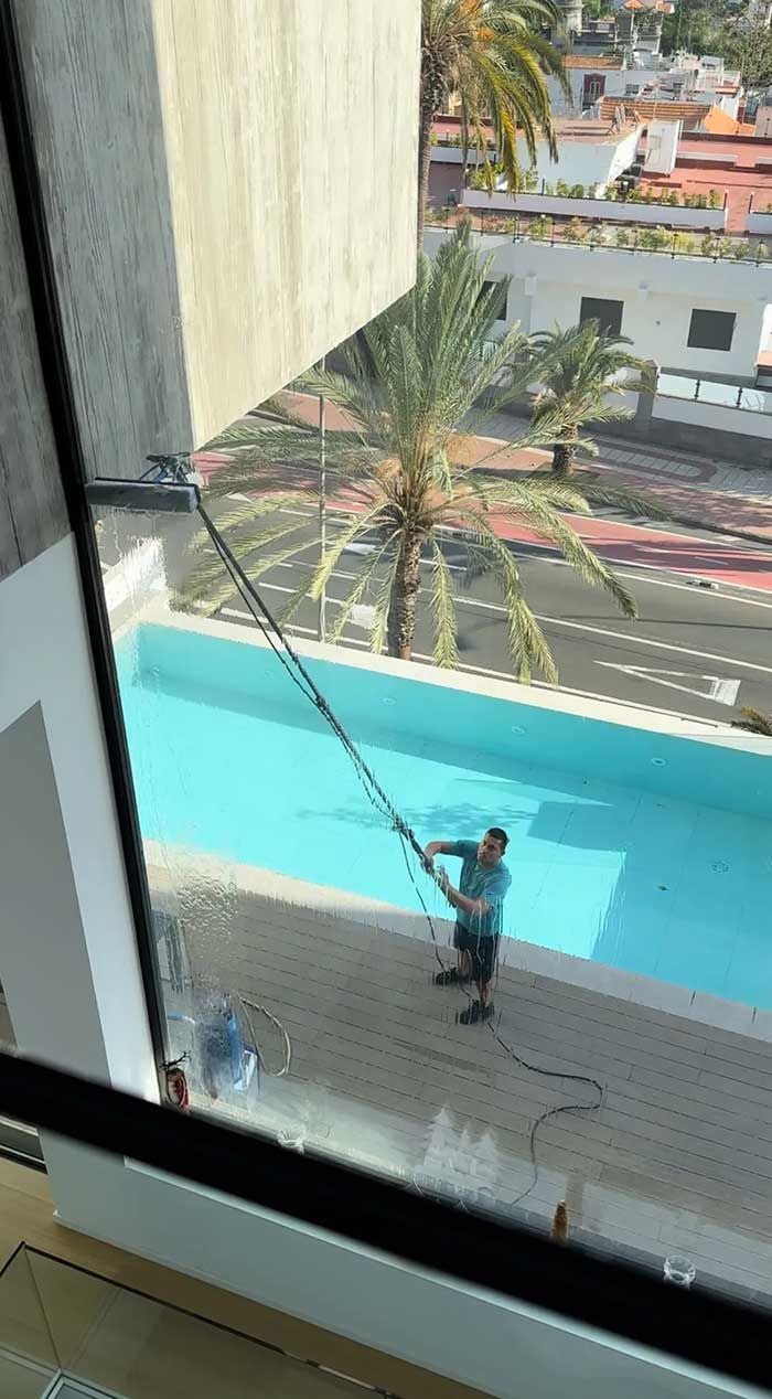 Una persona utiliza una herramienta de mango largo para limpiar una ventana exterior al lado de una piscina.