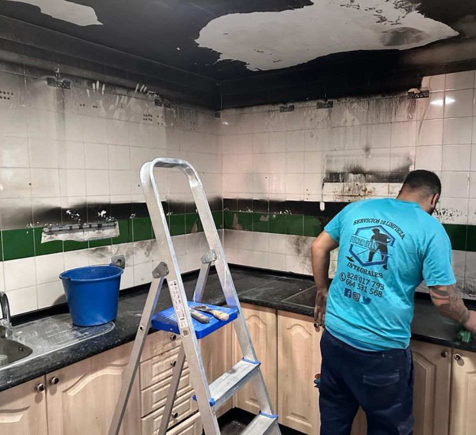 Hombre limpiando una cocina cubierta de hollín tras un incendio. Hay una escalera y un cubo. Azulejos negros y verdes.