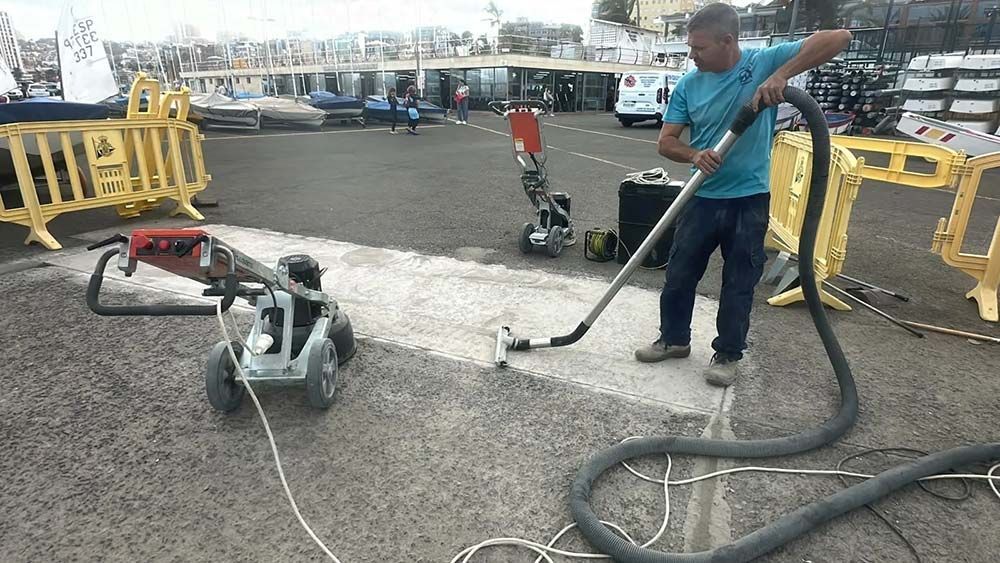 Hombre usando una aspiradora para limpiar una superficie pavimentada, posiblemente una carretera. Máquina gris cerca, barreras amarillas.