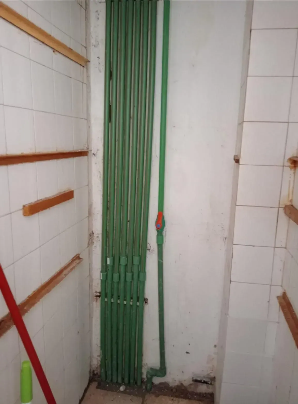 Tuberías de plomería verdes instaladas verticalmente contra una pared de azulejos blancos en un espacio de servicio.