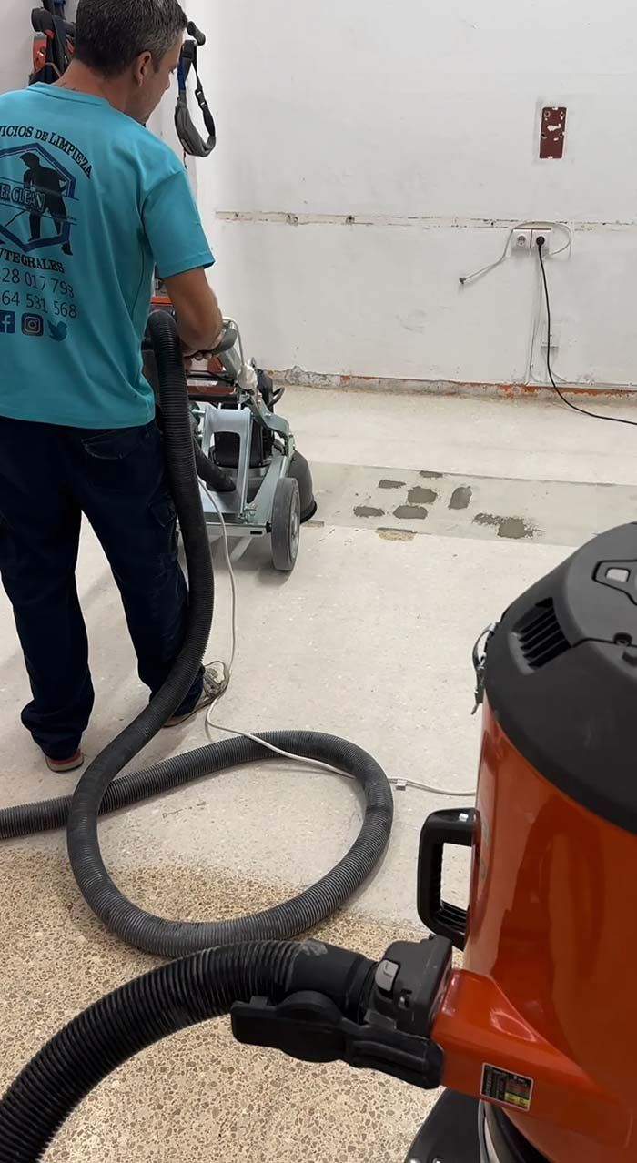 Persona puliendo un piso de concreto, conectada a una manguera de vacío en un espacio interior.