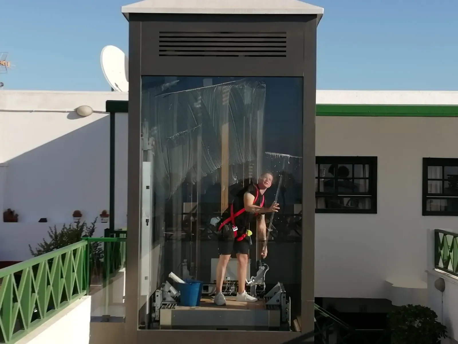 Una persona lava el cristal exterior de un pequeño ascensor. Lleva arnés de seguridad, de pie en el andén, cerca de un edificio blanco.
