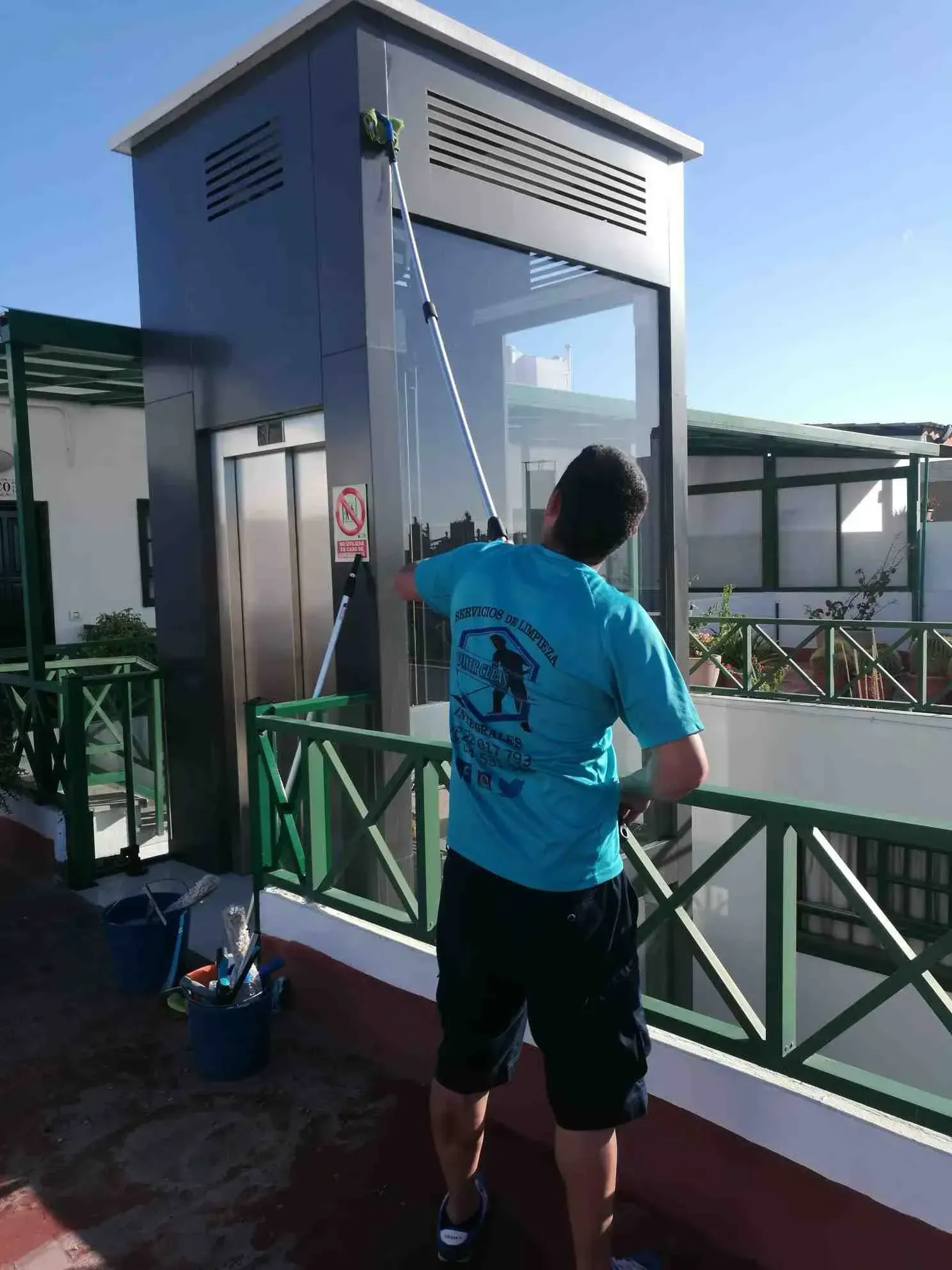 Hombre lavando la ventana exterior de un ascensor con una herramienta de mango largo en una azotea, en un día soleado.
