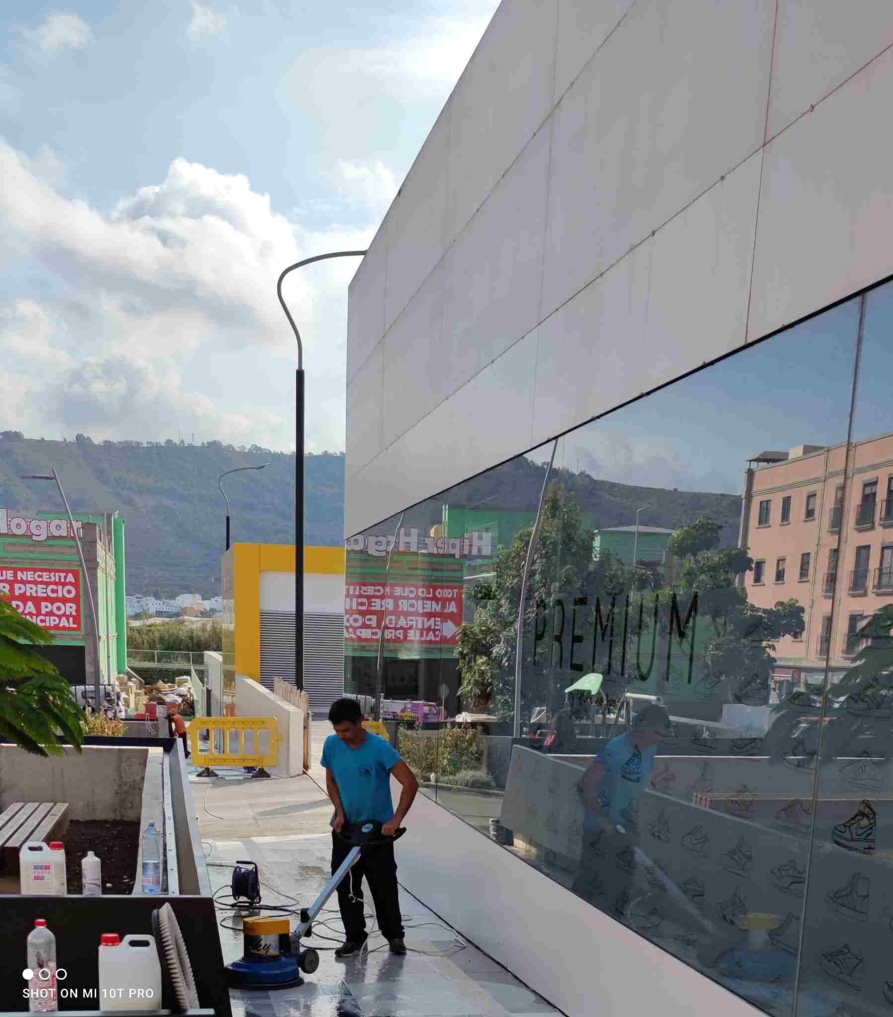 Hombre usando una pulidora de pisos afuera de un edificio. Pared de vidrio reflectante, cielo nublado y montañas.