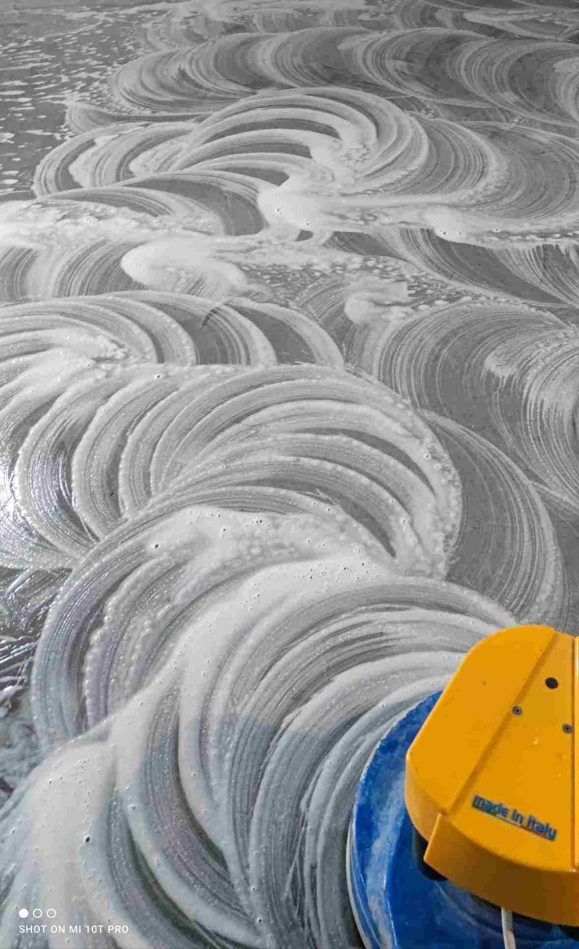 Piso limpiado con agua jabonosa, con patrones en espiral visibles. Fregadora amarilla y azul en primer plano.