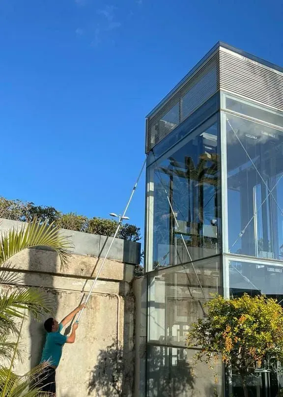 Persona limpiando un edificio de vidrio con una herramienta de mango largo bajo un cielo azul claro.